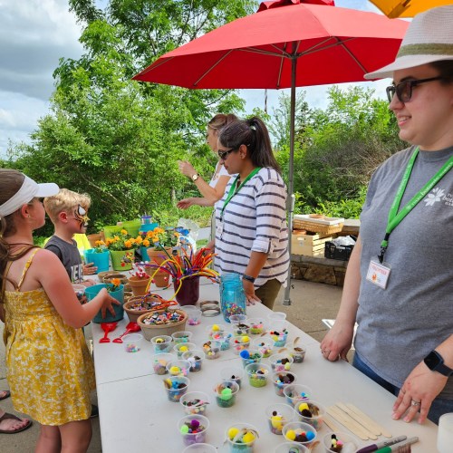 Children and adults engage in a craft activity at a table filled with colorful materials under umbrellas.