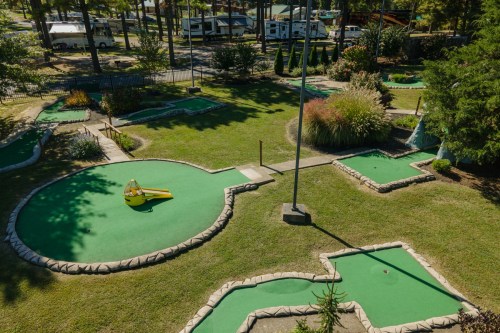 Aerial view of a mini-golf course with green turf, obstacles, and surrounding trees in a recreational area.