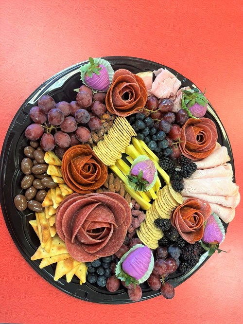 A colorful charcuterie platter with meats, cheeses, fruits, nuts, and decorative rose-shaped salami and cupcakes.