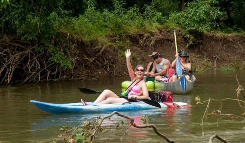 Three people kayaking on a calm river, with one woman waving and two others paddling in the background.