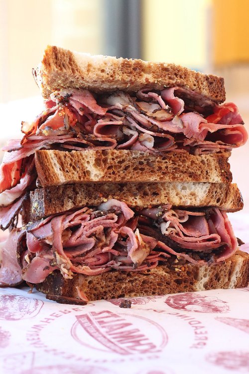 A towering sandwich stacked with layers of deli meats between slices of dark bread, resting on a patterned paper.