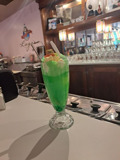 A vibrant green dessert drink topped with whipped cream and colorful sprinkles, served in a tall glass at a diner.
