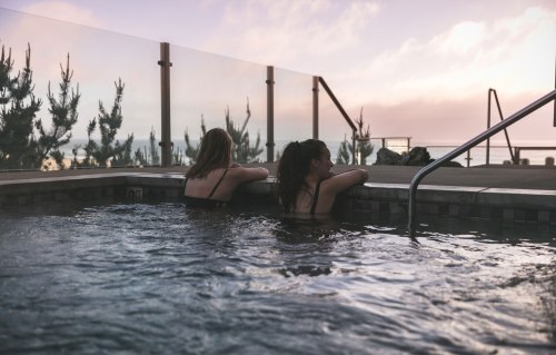 Two women relax in a hot tub at sunset, with a scenic view and trees in the background.