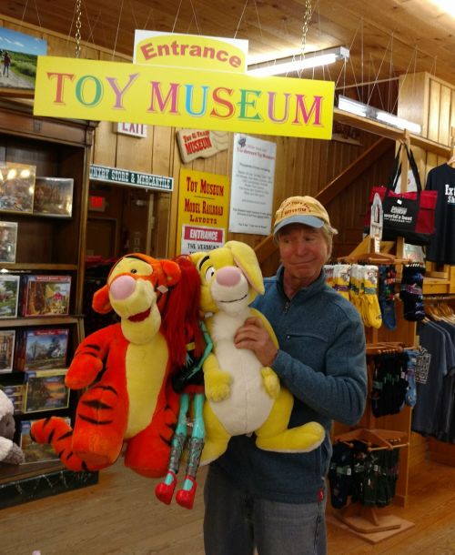 A man holds large plush toys of Tigger and a kangaroo in front of a "Toy Museum" entrance sign.