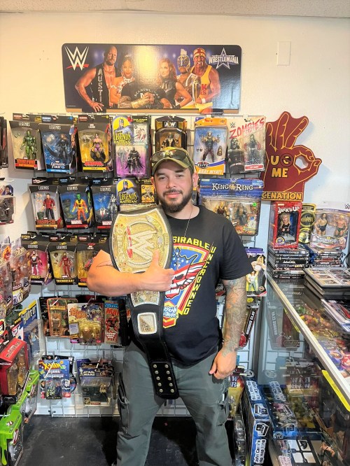 A man in a wrestling-themed shirt holds a championship belt in a store filled with action figures and collectibles.
