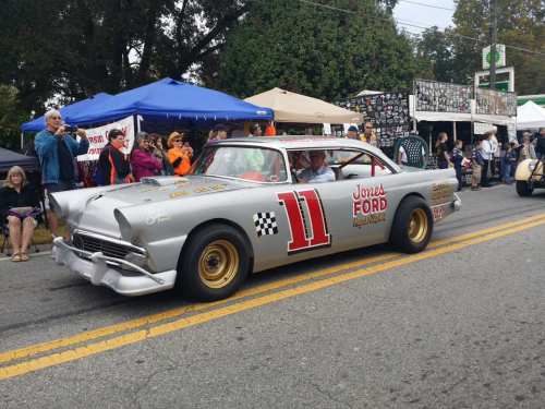 A vintage silver race car with the number 11 and "Jones Ford" branding drives past a crowd at a street event.