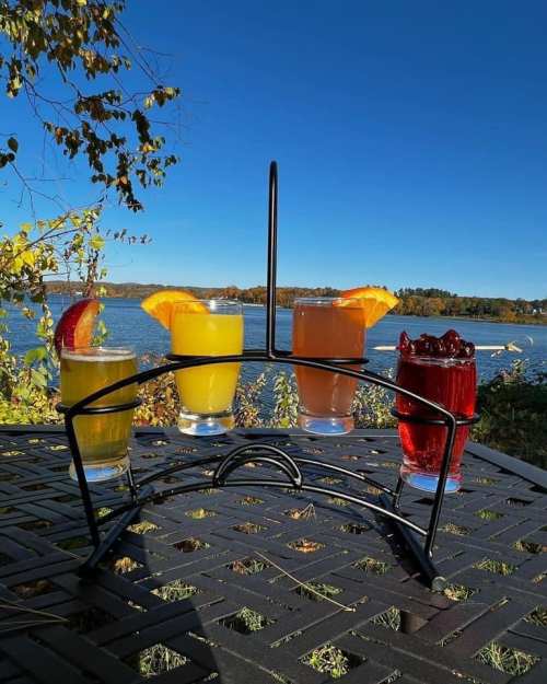 A display of four colorful drinks on a table by a lake, with a clear blue sky in the background.