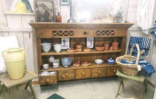 A rustic wooden shelf displays various pottery and kitchenware, flanked by two green chairs and a woven basket.