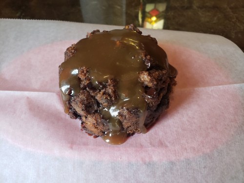 A round chocolate dessert topped with a glossy caramel sauce, resting on pink parchment paper.