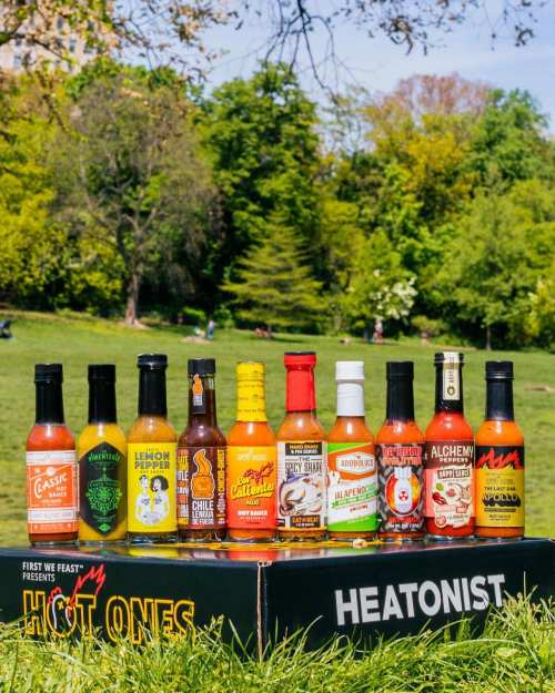 A lineup of colorful hot sauce bottles displayed on a grassy area, with trees and a park in the background.