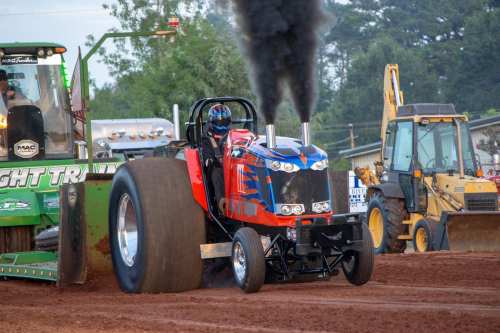 A colorful tractor pulling vehicle emits black smoke during a dirt track competition, with spectators and machinery in the background.