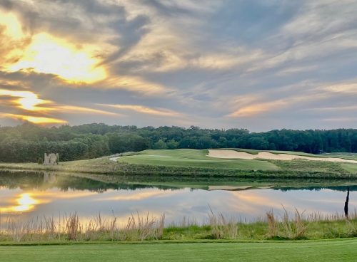 A serene landscape featuring a golf course at sunset, with reflections in a calm lake and lush greenery.