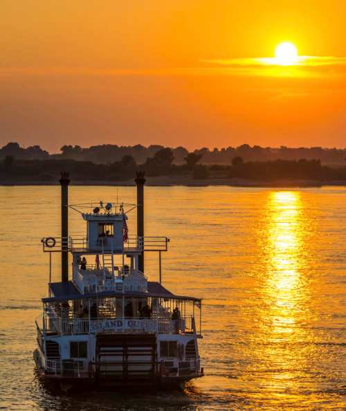 A steamboat glides on water at sunset, with vibrant orange and gold reflections and silhouettes of trees in the background.