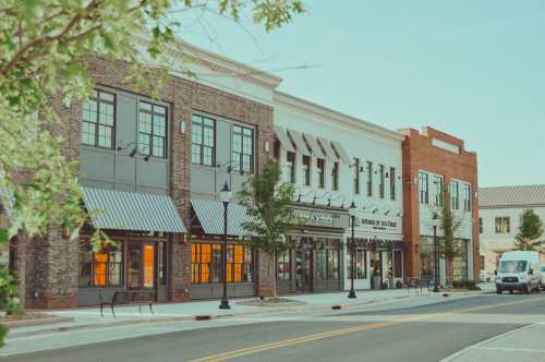 A charming street view of brick buildings with storefronts, trees, and a parked van under a clear sky.