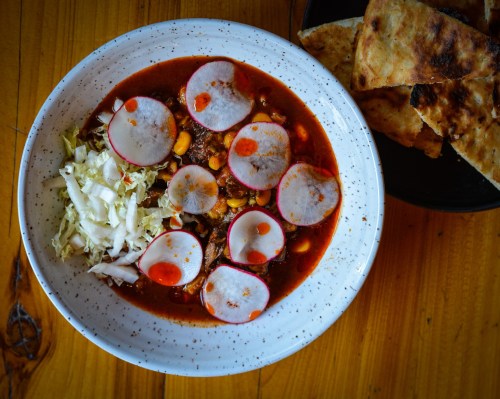 A bowl of spicy soup with radishes, corn, and cabbage, served with flatbread on the side.