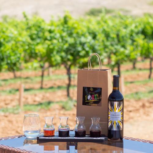 A bottle of wine and tasting glasses on a table, with a vineyard in the background under clear blue skies.