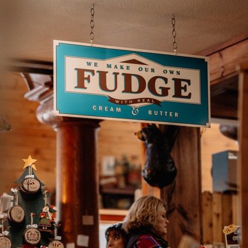 Sign in a shop reads "We Make Our Own Fudge" with a festive tree in the foreground and wooden decor in the background.