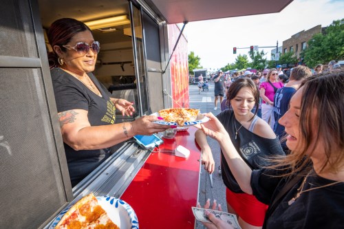 A food truck vendor hands a slice of pizza to a customer, while another customer waits in a busy outdoor setting.