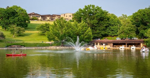 A serene lake scene with a fountain, surrounded by greenery and buildings, featuring kayakers and paddle boats.