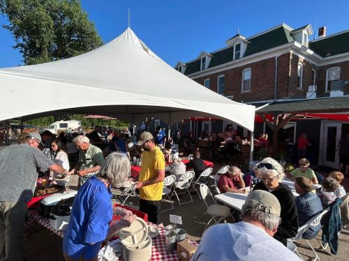 A lively outdoor gathering under a large tent, with people enjoying food and conversation at tables.