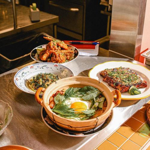 A variety of dishes on a table, including a clay pot with herbs, fried chicken, and two side dishes in decorative bowls.