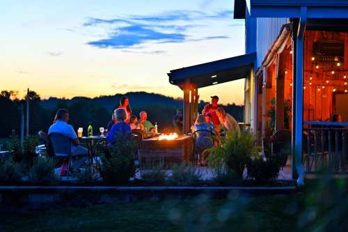A group of people gather around a fire pit at sunset, enjoying drinks and conversation near a cozy building.