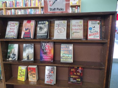 A wooden shelf displaying various "Staff Picks" books, featuring titles and authors like "The Silent Patient" and "You Love Me."