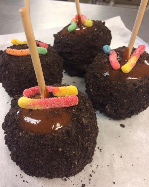 Four chocolate-covered treats coated in crushed cookies, topped with colorful gummy candies and wooden sticks.