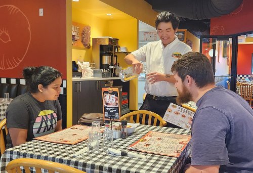 A waiter pours water for two diners at a restaurant with a checkered tablecloth and menus on the table.