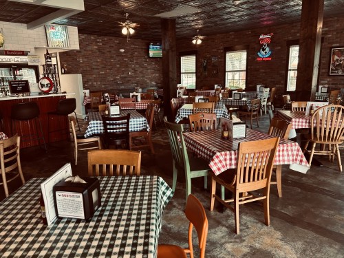 Interior of a casual restaurant with checkered tablecloths, wooden chairs, and a bar area. Bright and inviting atmosphere.