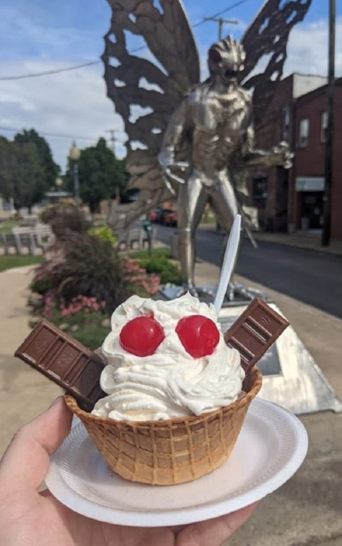 A hand holds a sundae in a waffle cone topped with whipped cream and cherries, with a butterfly statue in the background.