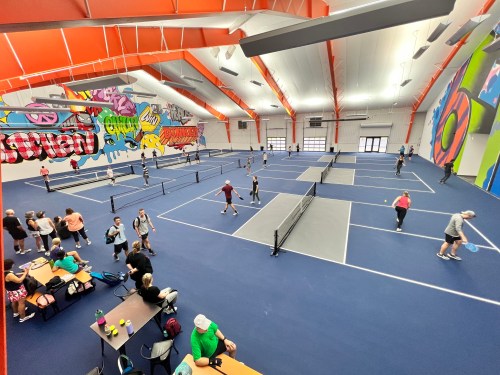A vibrant indoor tennis facility with multiple courts and players engaged in matches, surrounded by colorful murals.