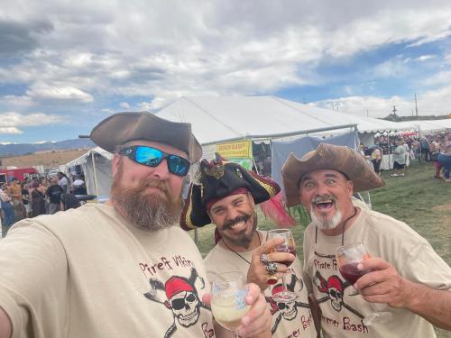 Three men in pirate hats and themed shirts hold drinks, smiling at the camera with a festival tent in the background.