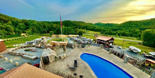 Aerial view of a pool area with stone features, surrounded by green hills and parked vehicles in a scenic landscape.
