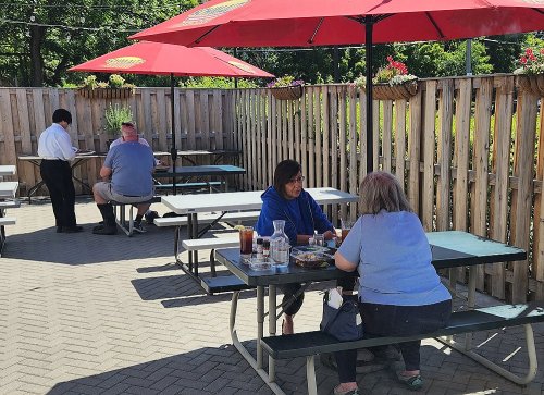 Two women sit at a table outdoors, enjoying a meal, while a waiter attends to another table under red umbrellas.