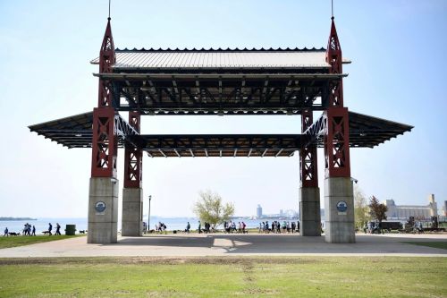 A large open pavilion with a metal frame overlooks a park and waterfront, with people walking in the background.