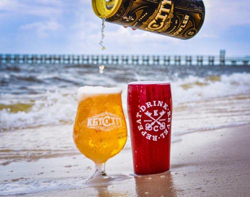 A beer can pours into a glass on a sandy beach, with waves and a pier in the background.