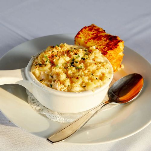 A bowl of creamy macaroni and cheese topped with breadcrumbs, served with a slice of cornbread on a white plate.