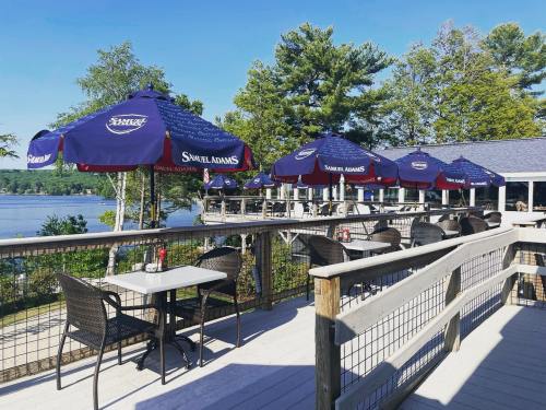 Outdoor dining area with tables and umbrellas overlooking a lake, surrounded by trees on a sunny day.