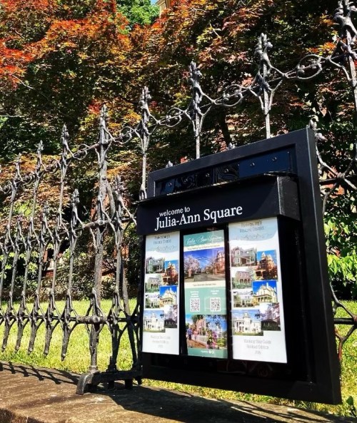 Sign welcoming visitors to Julia-Ann Square, featuring images and information about the area, set against a lush background.