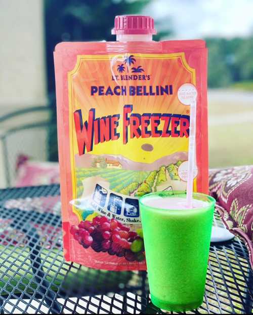 A colorful green drink in a cup next to a pouch of peach bellini wine freezer mix on a table outdoors.