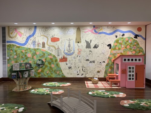 A colorful mural with whimsical illustrations, a playhouse, and green rugs in a children's play area.