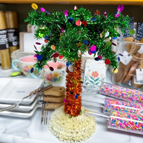 A colorful, sparkly mini palm tree with decorations, set on a table with various kitchen items in the background.