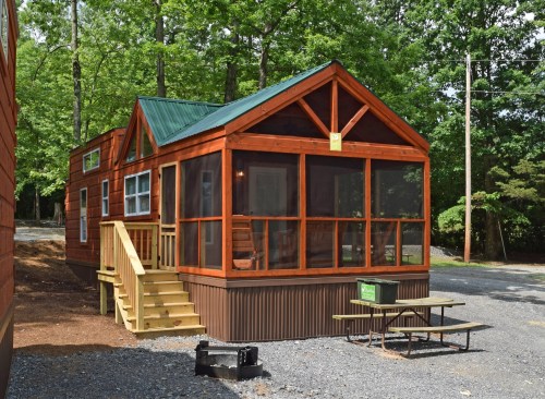 A cozy wooden cabin with a green roof, screened porch, and picnic table, surrounded by trees in a serene setting.