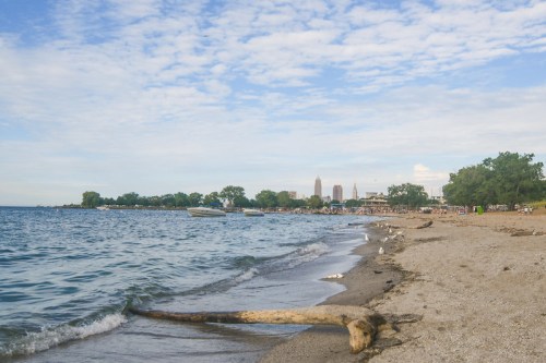 A sandy beach with gentle waves, boats in the water, and a city skyline in the background under a partly cloudy sky.