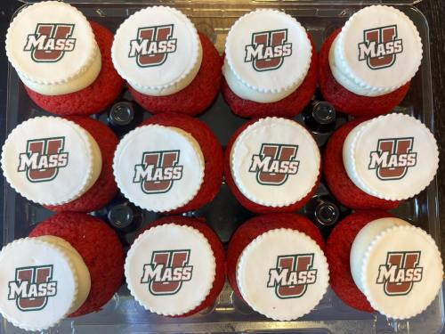 A dozen red velvet cupcakes topped with white frosting and UMass logos on circular fondant discs.