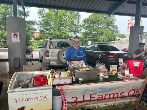 A vendor at a market stands behind a table with food, wearing sunglasses and a blue shirt, with cars in the background.