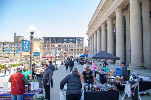 A bustling outdoor market with vendors and shoppers near a historic building on a sunny day.