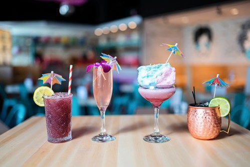 Four colorful cocktails with decorative umbrellas on a wooden table in a vibrant restaurant setting.