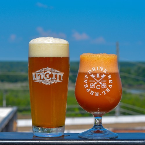 Two glasses of beer on a railing with a scenic background; one is a tall glass with a logo, the other a round glass.